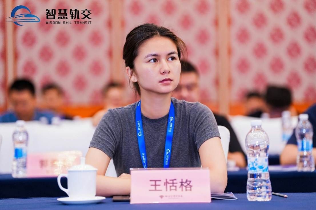 The image shows a person seated at a table with a blue tablecloth, a water bottle, and a notepad, amidst a group of people in a room with a decorative patterned wall. AI 生成的内容可能不正确。